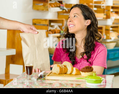 Pretty brunette woman wearing pink shirt sitting by table inside bakery, receiving brown paper bag from waiter and smiling happily Stock Photo