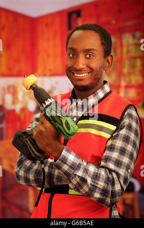 Young engineer carpenter wearing square pattern flanel shirt with red safety vest, holding handheld electric power drill smiling to camera Stock Photo