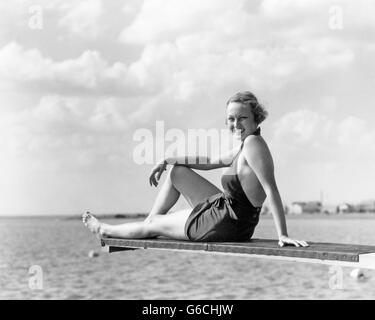 1930s SMILING WOMAN IN BATHING SUIT SITTING ON EDGE OF DIVING BOARD OVER WATER LOOKING AT CAMERA Stock Photo