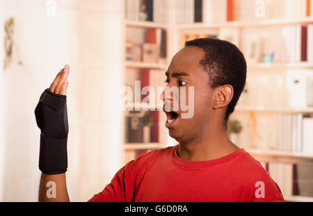 Man in red shirt staring at his own arm with shocked facial expression, wearing wrist brace support, blurry bookshelves background Stock Photo