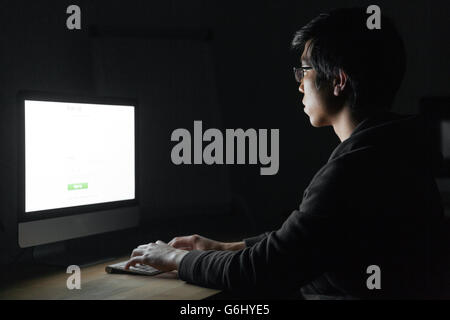 Focused asian young man in glasses working with computer in dark room Stock Photo
