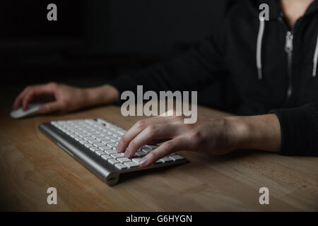 Hands of young man sitting at the table and typing on keyboard in dark room Stock Photo