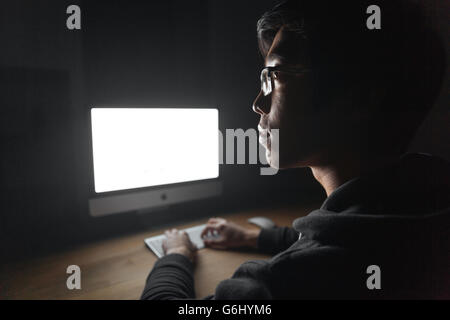 Thoughtful young man in glasses thinking and using blank screen computer in dark room Stock Photo