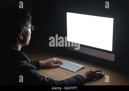Concentrated young man in glasses sitting at the table and using computer in dark room Stock Photo