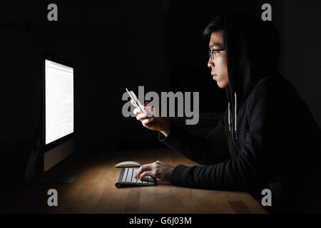 Serious young hacker using laptop and mobile phone in dark room Stock Photo