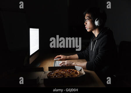 Man in headphones working with computer and eating pizza in dark room Stock Photo