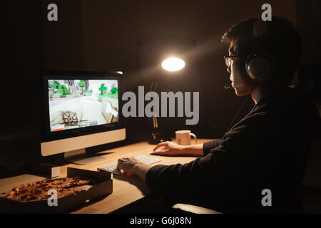 Man gamer in headset playing computer game and eating pizza in dark room Stock Photo