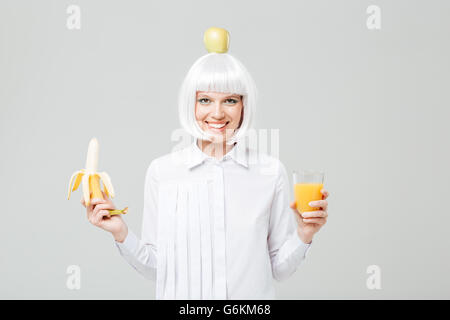 Cheerful young woman with apple on her head holding banana and glass of juice over white background Stock Photo