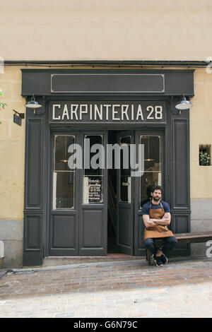 Craftsman with leather apron and arms crossed sitting at the entrance of his workshop Stock Photo