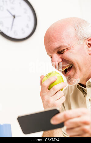 Single happy senior man getting ready to eat a green apple with phone in hand at work with clock near his head Stock Photo