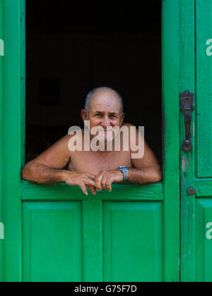 Cuban man, smiling friendly as he looks out of the green window door to a house in colonial Trinidad, Cuba Stock Photo
