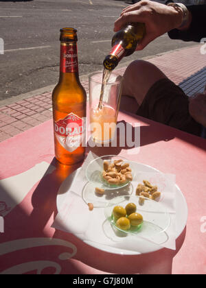 Sun shining through a brown Dorada bottle, mans hand pouring beer into half filled glass, nuts and olives, bar in Tenerife Spain Stock Photo