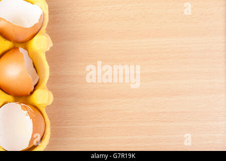 Above view of three broken egg shells in a yellow egg carton on the left hand side of a wooden background Stock Photo