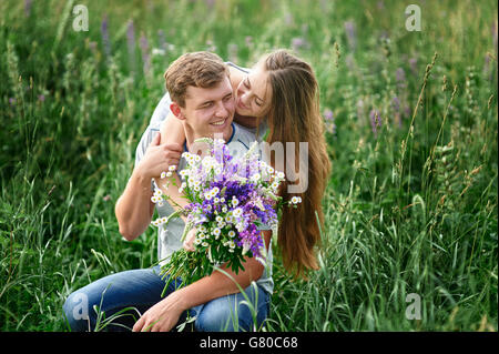 beautiful woman with bouquet of wild flowers hugging man sitting on the grass in meadow Stock Photo