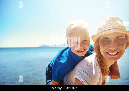 Happy little boy getting a piggy back ride from his attractive smiling mother in sunhat and sunglasses as they relax on a tropic