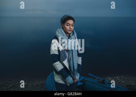 Teenage girl in warm clothing standing by inflatable raft on lakeshore Stock Photo
