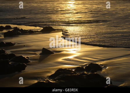 gentle waves wash over golden sand reflecting the sunrise on a pebble strewn shoreline on the Gower Stock Photo