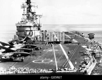 Sailors bask in the sun on board the aircraft carrier HMS Ark Royal in the Mozambique Channel, while Gannet and Sea Vixen aircraft are airborne. Other aircraft stand ready on deck for the next sortie. Stock Photo