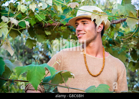 Side view of handsome young man in hat toching vine leaves in garden in sunlight Stock Photo