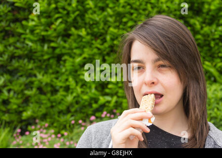 Young woman, adult or late teens, headshot in a park eating a cereal bar. Stock Photo