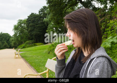 Young  woman, adult or late teens, seated in park eating a slice of apple. Stock Photo