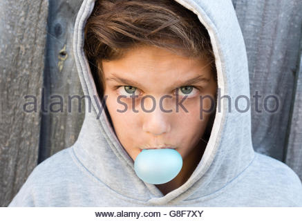 Portrait of aggressive teenage boy with hoodie blowing bubble gum against grey wooden fence