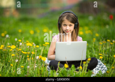 Little girl in headphones with laptop sitting on the grass in the Park. Stock Photo