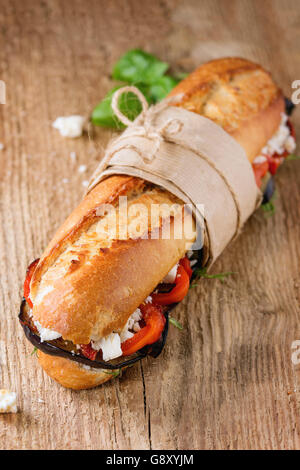 Papered vegetarian baguette submarine sandwich with grilled eggplant, pepper and feta cheese on wooden chopping board over dark Stock Photo