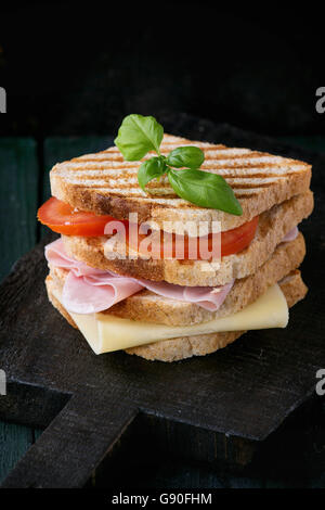 Whole grain grilled sandwich bread with ham, cheese and tomatoes on black wooden chopping board over dark background. Stock Photo