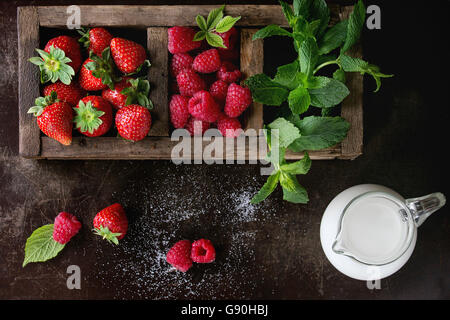 Fresh berries strawberry and raspberry with bunch of mint in old wooden sectioned box and jug of milk over dark textured backgro Stock Photo