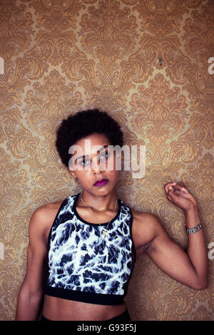 Beauty in dereliction : Fashion photography of a young afro-caribbean woman girl alone in an ornately  wall-papered room  wearing fitness-style clothes, looking sultry and moody