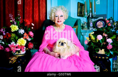 Portrait of novelist and author Barbara Cartland at home with one of her beloved dogs, sitting in one of the gaily painted rooms in a bright pink dress offset by the flowers that surround her and the peacock blue walls.  Dame Barbara lived and worked since 1950 at Camfield Place, formerly the home of Beatrix Potter, a ten-bedroom mansion set in 400 acres of farmland and woods near Hatfield, Hertfordshire. Stock Photo