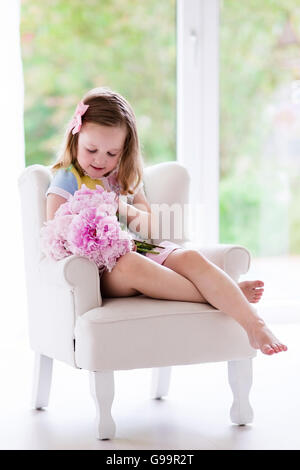 Little girl smelling peony flower bouquet sitting in a white chair in sunny bedroom. Nursery interior with flowers for girls Stock Photo