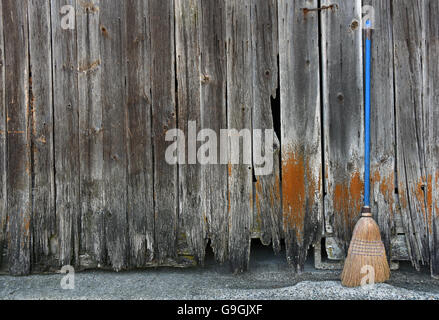 Old worn broom with blue handle leaning against weathered wooden barn. Stock Photo