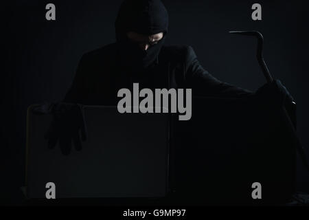 Angry computer hacker in suit stealing data from laptop with crowbar and gloves in front of black background Stock Photo