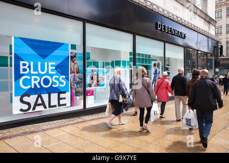 Out of focus blurred shoppers, people walking figures passing Blue Cross Summer Sale signs at Debenhams Store in Piccadilly, Manchester, UK. Sale and reductions, deals & offers, clearance items, discount, price, promotion, retail business, Everyday lifestyle street scenes outside Debenhams department store. Stock Photo
