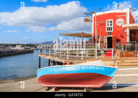PUERTO DEL CARMENT PORT, LANZAROTE - JAN 17, 2015: colorful fishing boat in front of red restaurant building in Puerte del Carmen port. This town is popular holiday destination on Lanzarote island. Stock Photo