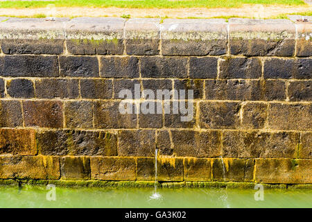 Wet stone blocks at a canal wall with some water flowing out of a small hole in the wall. Stock Photo