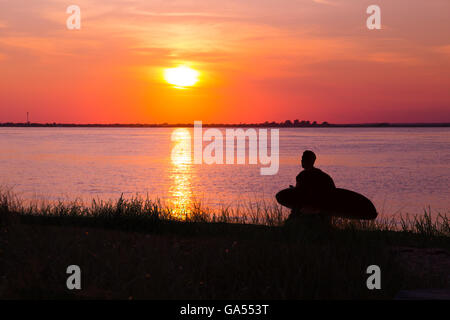 Silhouetted surfer with board walking along shore at sunset Stock Photo