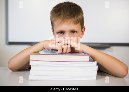 Portrait of sad schoolboy leaning on stack of books in class room Stock Photo