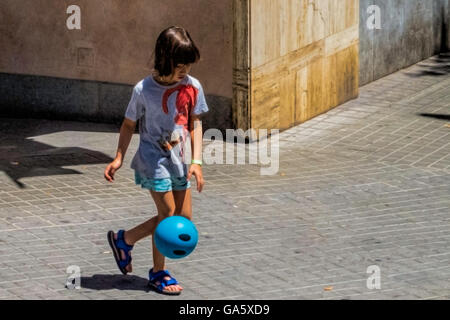 A ten year old girl, in shorts and sandals, playing with a blue ball on a city street. Stock Photo