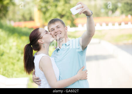 Beautiful young loving couple standing in park embracing. Pretty casual woman kissing her handsome man. He is holding a phone Stock Photo