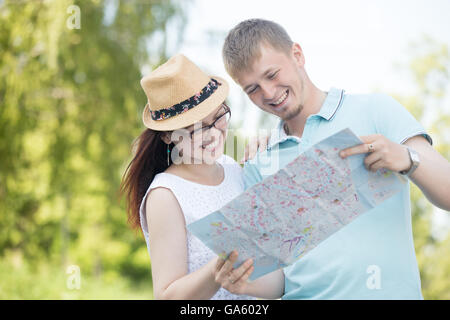 Cheerful young traveling beautiful couple standing in street on summer day looking at map and smiling. Pretty woman wearing Stock Photo