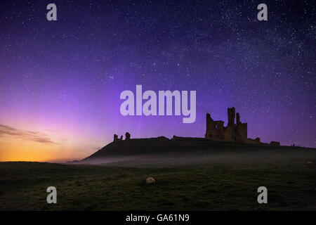 Dunstanburgh Castle at night with low mist and the northern lights dancing across the sky. Stock Photo