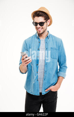 Cheerful young man in sunglasses and hat standing and using smartphone Stock Photo