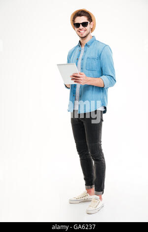 Smiling young man in sunglasses and hat holding and using tablet over white background Stock Photo