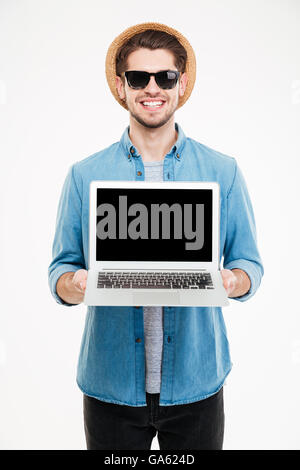 Smiling young man in sunglasses and hat standing and holding blank screen laptop Stock Photo
