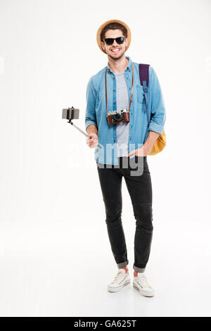 Happy young tourist holding backpack, vintage camera and smartphone on selfie stick over white background Stock Photo