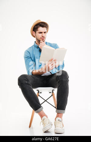 Concentrated handsome young man in hat sitting and reading a book over white background Stock Photo