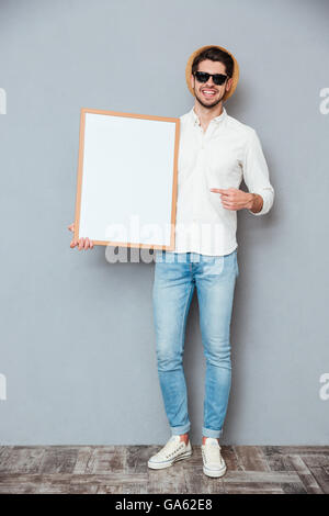 Cheerful young man holding blank white board and pointing on it over grey background Stock Photo